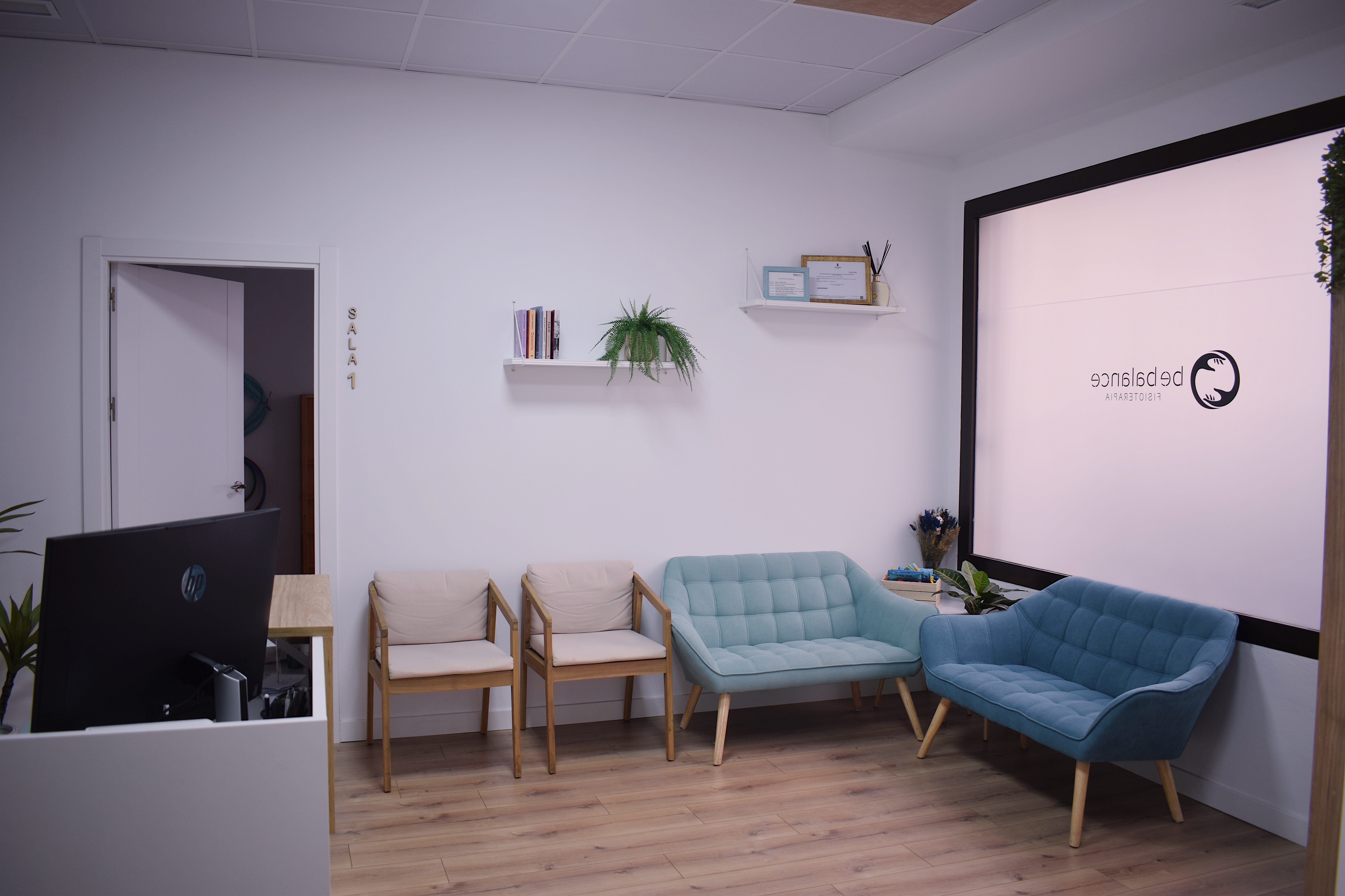 Sala de espera de Be Balance Fisioterapia, con sillas, dos sofás, mostrador de recepción y el logotipo en el cristal