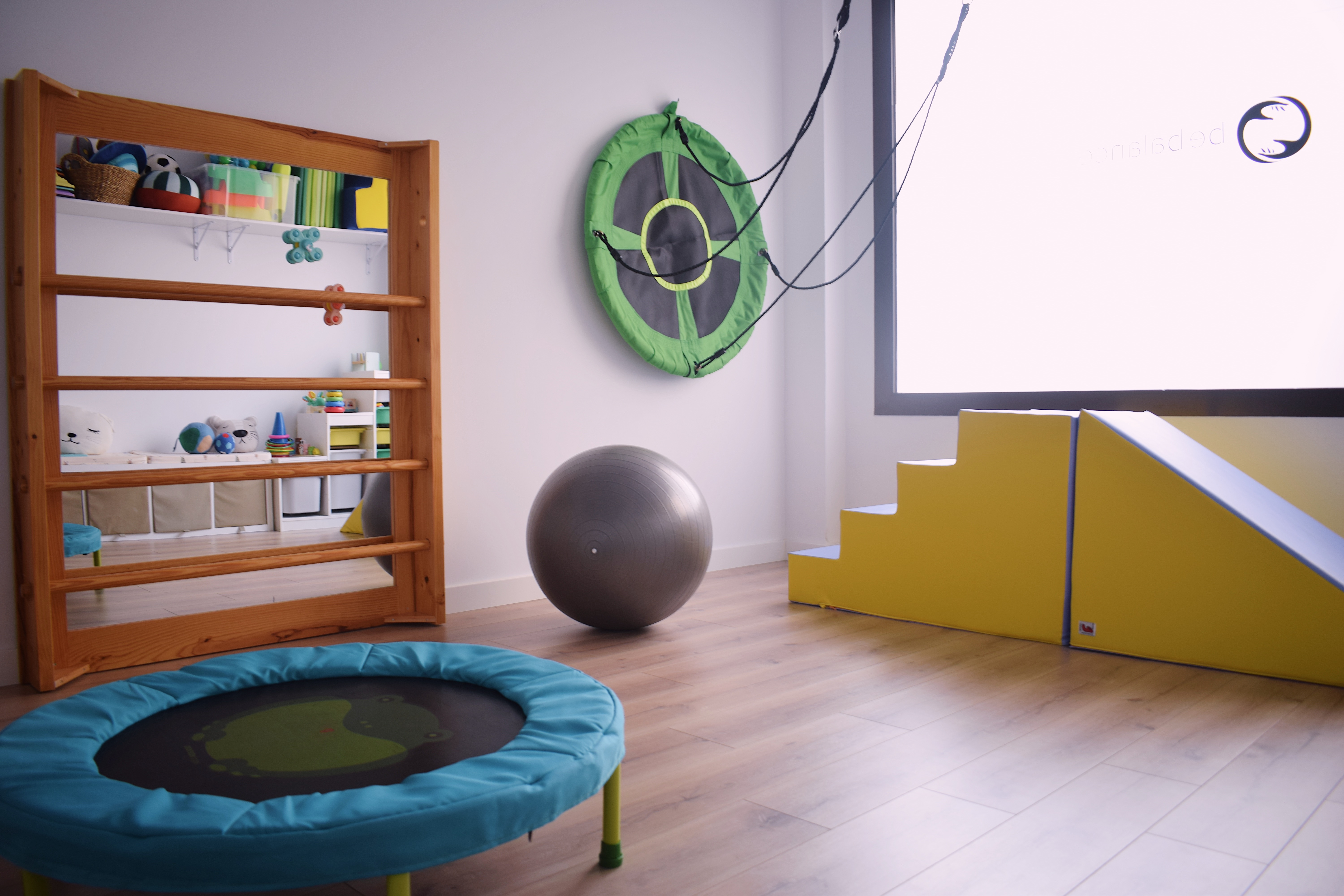 Sala de fisioterapia pediátrica con espaldera, cama elástica, columpio, pelota grande y rampa de espuma